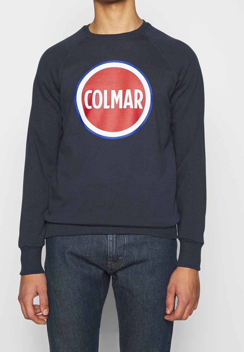 Mörkblå sweatshirt med runt rött logo som har "COLMAR" i vitt, ribbad mudd och nederkant, mjukt bomullsmaterial, avslappnad passform.