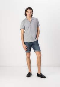 Chemise à manches courtes gris clair à bouton, associée à un short en denim retroussé. Le modèle porte des mocassins noirs. Texture lisse, coupe décontractée.