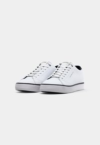 Paire de baskets basses blanches avec semelles grises, lacets blancs et doublure intérieure noire, avec le texte Tommy Hilfiger sur le côté et la languette.