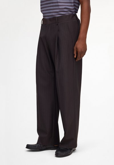 Man wearing black pinstripe wide-leg trousers with black loafers and a short-sleeve striped shirt, standing against a white background.