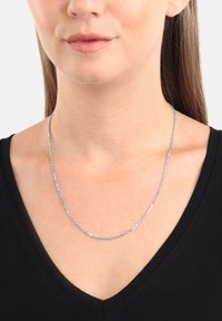 Silverkedja hängande på en kvinna som bär en svart V-ringad topp, som visar hennes hals och nedre del av ansiktet mot en neutral bakgrund.