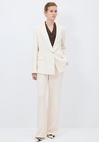 Costume crème à rayures avec un blazer ajusté à un seul bouton doré et un pantalon ample. Chemise à boutons marron en dessous.