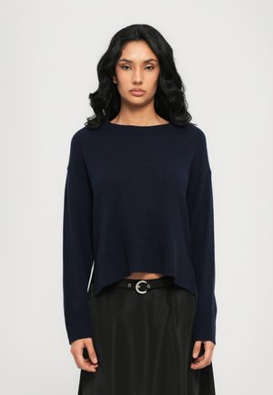 Navyblauer Strickpullover mit gerippten Bündchen und Saum, in lockerer Passform und mit überschnittenen Schultern, kombiniert mit einem schwarzen Rock und einem runden Gürtel.