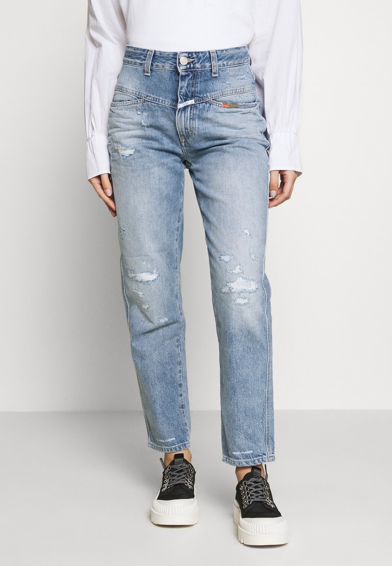 Personne portant un jean bleu clair taille haute et usé, une chemise blanche à manches longues et des baskets noires à semelle blanche épaisse.