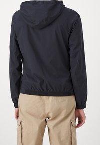 Man wearing a black hooded jacket and beige cargo pants, shown from the back against a plain white background.