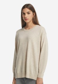 Hellbeige, oversized Pullover aus weichem Strickmaterial, mit geripptem Hals- und Armabschluss, lockerer Passform, minimalistische Details, kombiniert mit grauen Jeans.