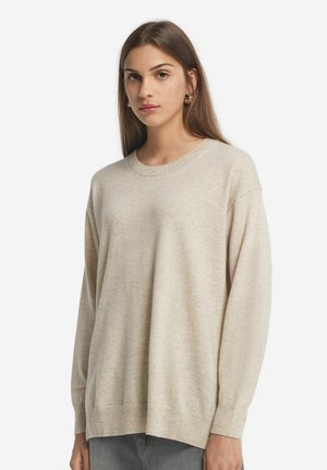 Maglione oversized beige chiaro, in morbido materiale knit, con scollatura e polsini a costine, vestibilità rilassata, con dettagli minimi, abbinato a jeans grigi.