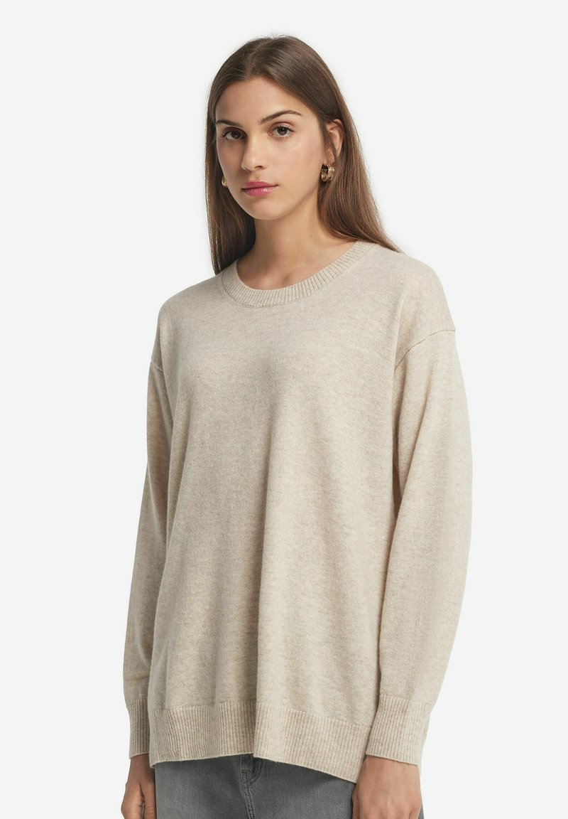 Hellbeige, oversized Pullover aus weichem Strickmaterial, mit geripptem Hals- und Armabschluss, lockerer Passform, minimalistische Details, kombiniert mit grauen Jeans.