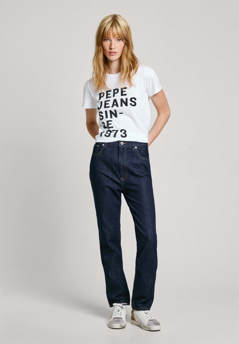 Weißes T-Shirt mit schwarzem Text "PEPE JEANS SINCE 1973," kombiniert mit dunkelblauen Jeans. Model trägt weiße Sneaker und steht vor einem grauen Hintergrund.