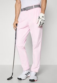 Růžové golfové kalhoty s hladkým povrchem, rovného střihu a šedým opaskem. Nosí se s golfovou rukavicí a bílými botami s černými akcenty.