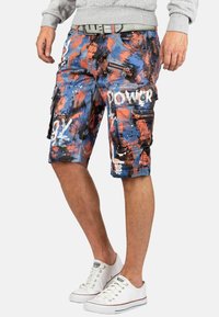 Shorts cargo en bleu, rouge et blanc avec un motif abstrait peint. Comprend plusieurs poches et zips. Associé à des baskets blanches.