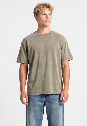 Giovane uomo con capelli chiari e ricci che indossa una maglietta oversize verde oliva sbiadita e jeans blu, che guarda leggermente a destra su uno sfondo semplice.
