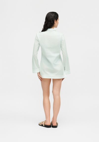 Γυναίκα με σκουρόχρωμα μαλλιά που φοράει λευκό μίνι φόρεμα με μακριά μανίκια και μπεζ σανδάλια, στέκεται με την πλάτη προς το μέρος μας μπροστά σε ένα απλό φόντο.