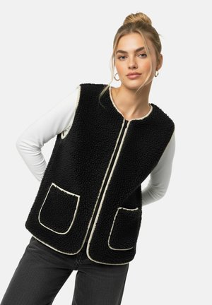 Femme portant un gilet polaire noir avec des bordures blanches et deux poches avant, par-dessus un haut blanc à manches longues, posant devant un fond uni.