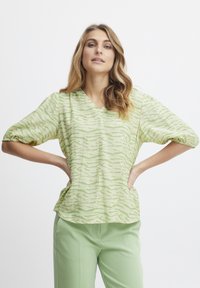 Blusa verde e crema con scollatura a V, caratterizzata da un motivo a onda testurizzato e maniche corte a sbuffo, abbinata a pantaloni verde chiaro.