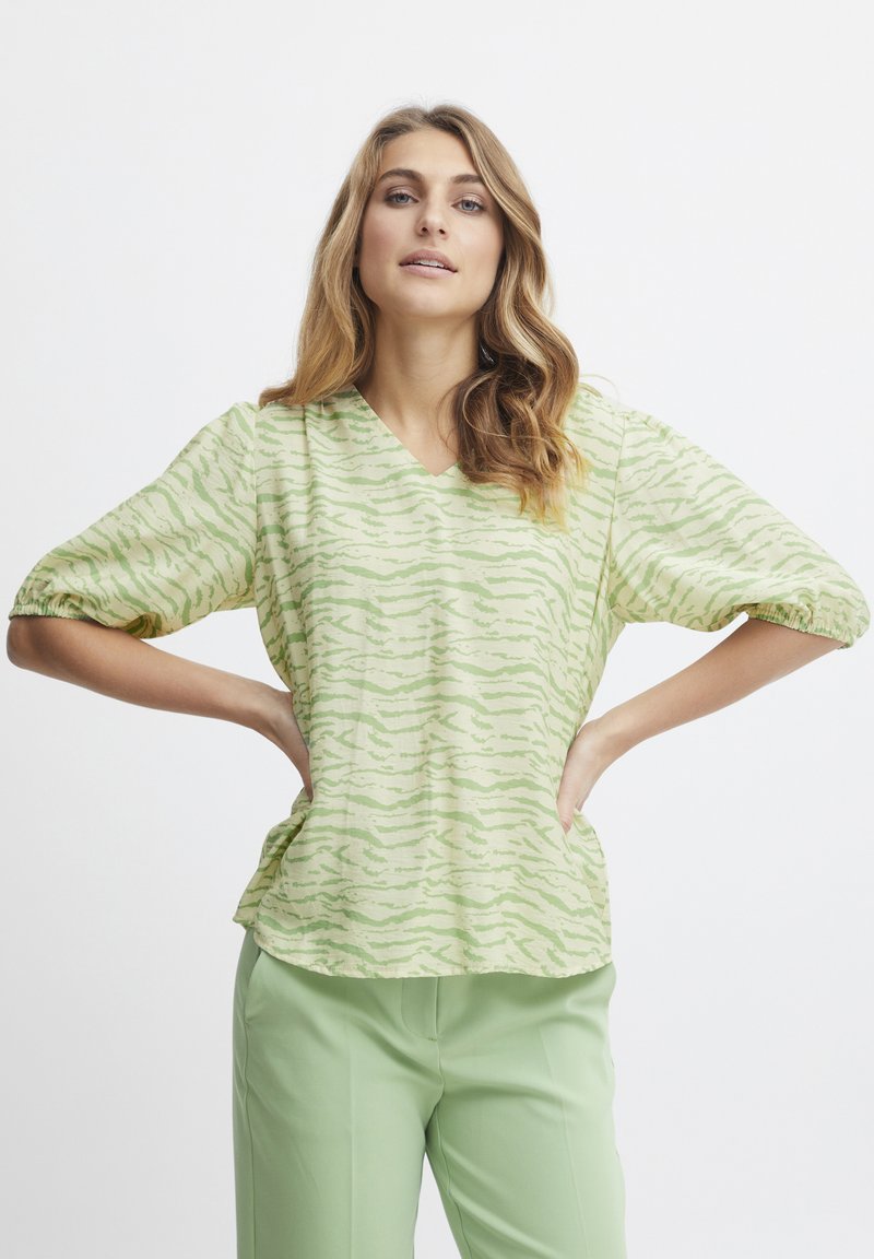 Blusa verde e crema con scollatura a V, caratterizzata da un motivo a onda testurizzato e maniche corte a sbuffo, abbinata a pantaloni verde chiaro.
