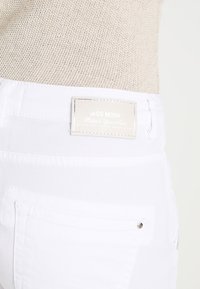 Jeans de mezclilla blanca con un cinturón texturizado, que presenta una etiqueta de logo y un bolsillo trasero con detalles de hardware plateado.