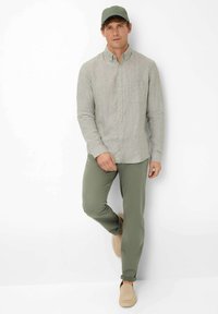 Light gray linen button-up shirt, olive green pants, and beige slip-on shoes. Model wearing a green cap, standing against a white background.