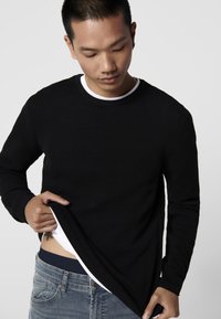 Maglietta nera a maniche lunghe con un collo tondo, indossata sopra una camicia bianca, abbinata a jeans azzurri chiari.