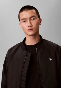 Jonge man met kort haar draagt een zwart Calvin Klein bomberjack en een zwart shirt, kijkt recht vooruit tegen een grijze achtergrond.