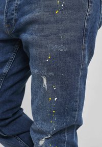 Tmavě modré džíny z denimu s opotřebovaným vzhledem, zdobené žlutými a bílými skvrnami na přední části stehen a ohrnutými detaily.