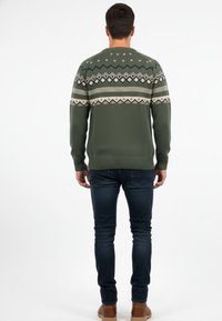 Jersey de punto verde oliva con patrones geométricos en crema y azul marino. Cuello redondo y puños acanalados; combinado con vaqueros oscuros y botas color marrón claro.