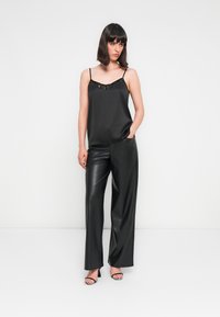 Femme portant un caraco noir à sequins, un pantalon en cuir noir à jambes larges et des sandales à talons noires et blanches à rayures, debout sur un sol blanc.