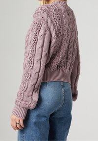 Stickad lila tröja med kabelmönster, ribbade manschetter och nederkant samt en lätt kort design, tillsammans med högmidjade blåjeans.