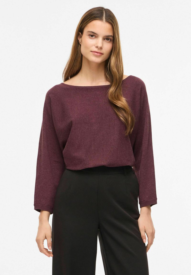 Jeune femme aux longs cheveux bruns portant un pull ample bordeaux et un pantalon noir taille haute, debout devant un fond clair.