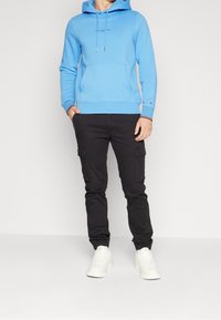 Felpa con cappuccio blu in cotone con tasca frontale e stringhe, abbinata a pantaloni cargo neri con tasche laterali e sneakers bianche.