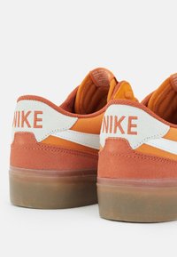 Sneaker in suede arancione con dettagli bianchi, dotate di un tallone texture, suola in gomma e branding stampato sulla parte posteriore del tallone.