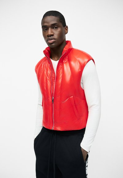 Άνδρας με φωτεινό κόκκινο αμάνικο γιλέκο πάνω από λευκό πουκάμισο με μακριά μανίκια και μαύρο παντελόνι, στέκεται μπροστά σε απλό φόντο.