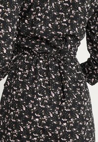 Mustassa kankaassa oleva mekko, jossa on vaaleanpunaisia ja vihreitä kukkakuoseja, vyötärö on kireä ja hihat puffatut. Materiaali on teksturoitu ja kevyt.