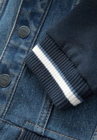 Gros plan sur une veste en denim avec un poignet côtelé bleu marine arborant des rayures blanches et bleu clair ainsi que des boutons noirs étiquetés "Authentic 1982 Next Dude".