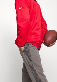 Haut rouge à manches longues Nike avec un logo blanc, associé à un pantalon gris présentant un motif noir texturé et des poches latérales, tenant un ballon de football.