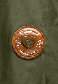 Brun läderpatch med en hjärtformad brassembel och vit sömnad, med texten "Raquar sedan 1997" på en grön tygbakgrund.