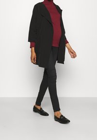 Abrigo negro sobre un cuello de tortuga a rayas burdeos, combinado con jeans ajustados negros y mocasines negros. Tejido suave y silueta relajada.