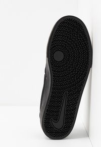 Semelle de chaussure noire avec un motif de bande de roulement en zigzag et un grand logo Nike swoosh près du talon, incliné contre un fond d'angle blanc.