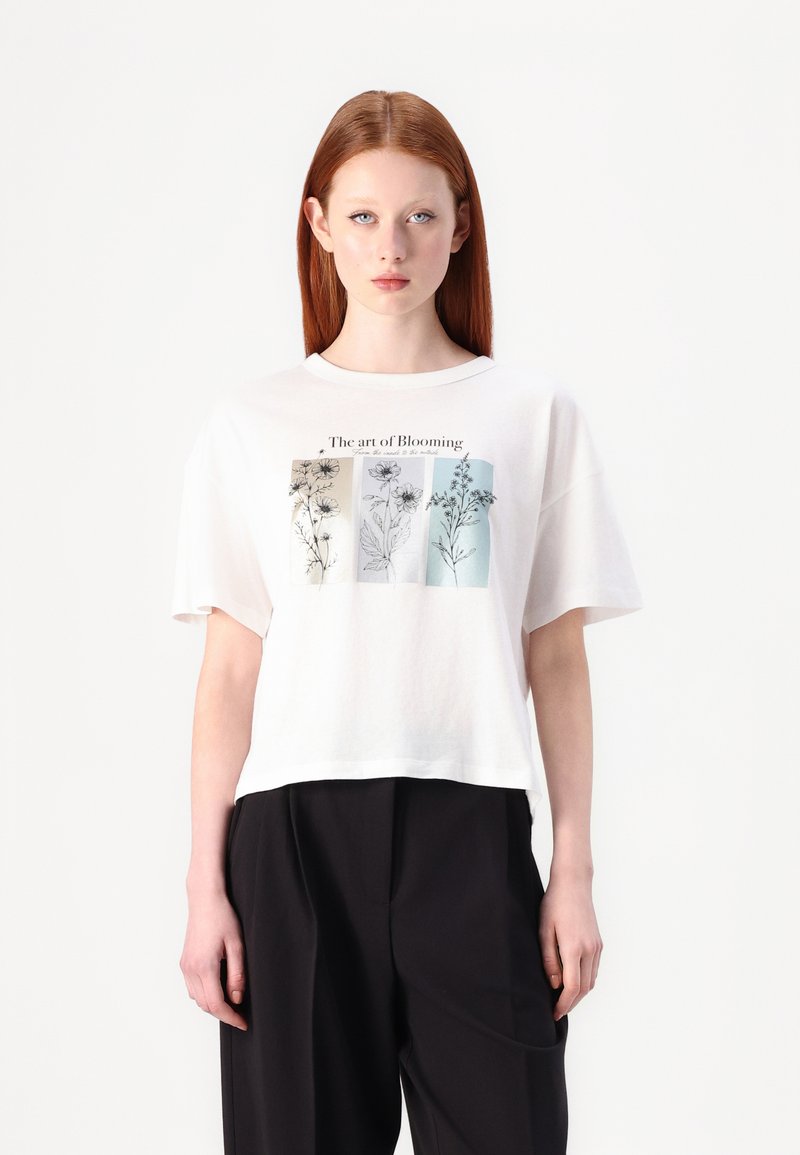 T-shirt en coton blanc à manches courtes, ornée d'un imprimé botanique dans des couleurs atténuées et du texte "L'art de Fleurir" en haut.