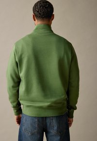 Green sweatshirt with a high collar and ribbed cuffs, made of soft fabric. Worn with blue denim jeans, focusing on the back view.