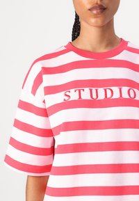 Sarkans un balts svītrainais T-krekls ar īsām piedurknēm, apaļu kakla izgriezumu un izšūtu "STUDIO" detaļu priekšpusē.
