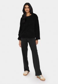 Crni teksturirani pulover s okruglim izrezom, uparen s crnim hlačama ravnog kroja i krem-bijelim tenisicama s zelenim akcentima.