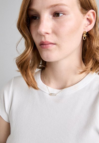 Pendientes de aro de oro y un delicado collar de cadena de oro con un colgante ovalado, llevados con una camiseta blanca de canalé. El cabello texturizado cae suavemente.
