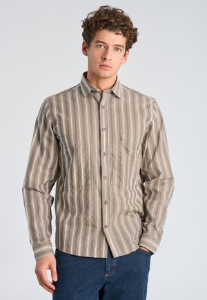 Junger Mann mit lockigem Haar, der ein beiges und weiß gestreiftes Hemd mit langen Ärmeln und Knöpfen sowie eine blaue Jeans trägt, steht mit einer Hand in der Tasche.