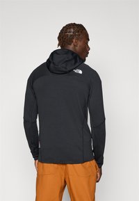 Veste à capuche noire en tissu texturé, avec des manches longues, un logo au dos, et un pantalon orange contrastant.