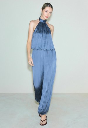 Kobieta ubrana w plisowany satynowy niebieski top na ramiączkach z dopasowanymi luźnymi spodniami i ciemnobrązowymi sandałami, pozuje przed jasnoszarym murem.