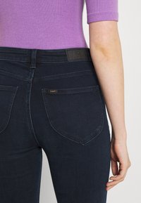 Jeans ajustados de color azul oscuro con una textura suave, que presentan bolsillos traseros y una etiqueta de cuero con la marca en la cinturilla.