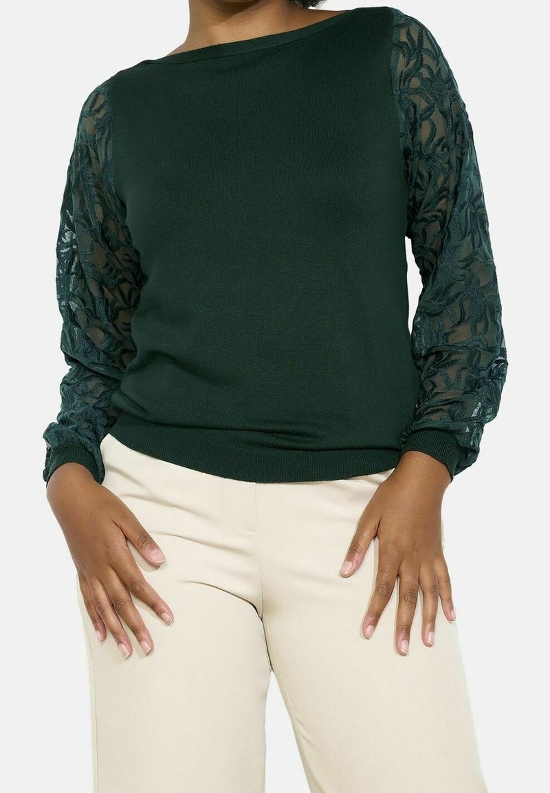 Femme portant un haut à manches longues vert foncé avec manches en dentelle florale transparente et pantalon beige clair, les mains reposant près des cuisses.