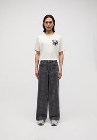 Cream cotton t-shirt with black floral design on the left chest, paired with wide-leg gray jeans featuring a subtle floral pattern.