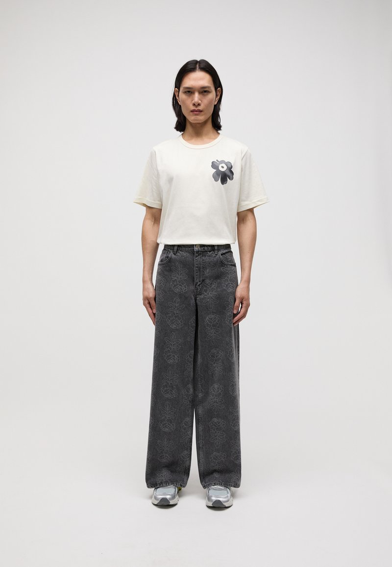 Cream cotton t-shirt with black floral design on the left chest, paired with wide-leg gray jeans featuring a subtle floral pattern.